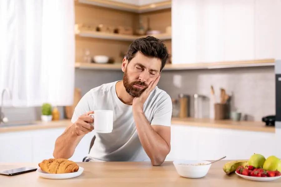 Unavený muž s hrnkem kávy podpírá hlavu u snídaně v kuchyni.