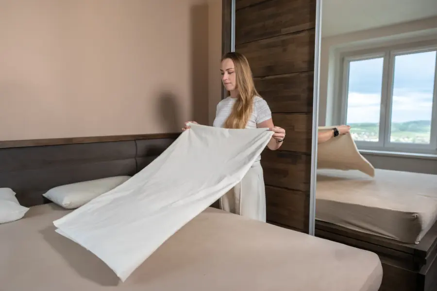 Žena v ložnici rozkládá nad postelí čisté bílé prostěradlo.